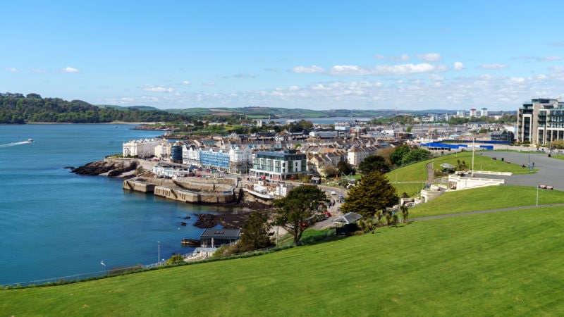 view from the grassy lawn at Plymouth Hoe looking towards a small area of the city above a grey sea wall with the sea to the left on a sunny day with blue sky. 