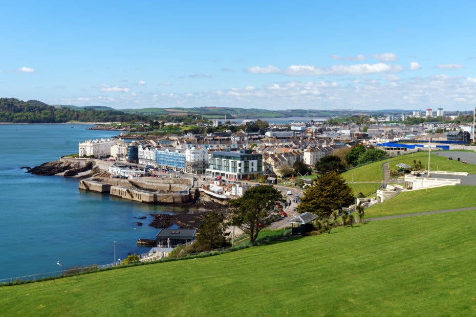 view from the grassy lawn at Plymouth Hoe looking towards a small area of the city above a grey sea wall with the sea to the left on a sunny day with blue sky.