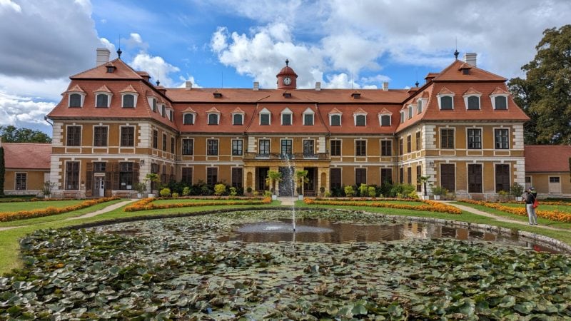 Large pong full of green waterlily leaves in front of a yellow chateau with red roof on a sunny day with a blue sky behind.