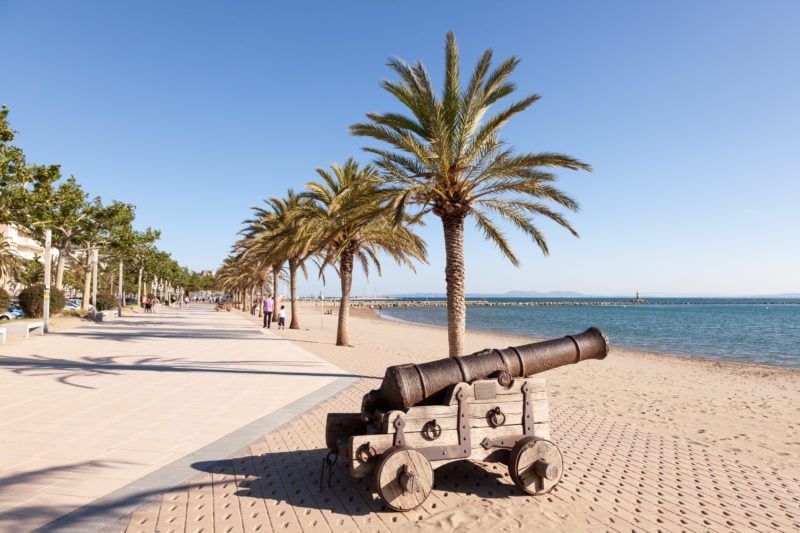 canon on a stone promenade next to a white sandy beach with a row of palm trees in roses on the costa brava in spain