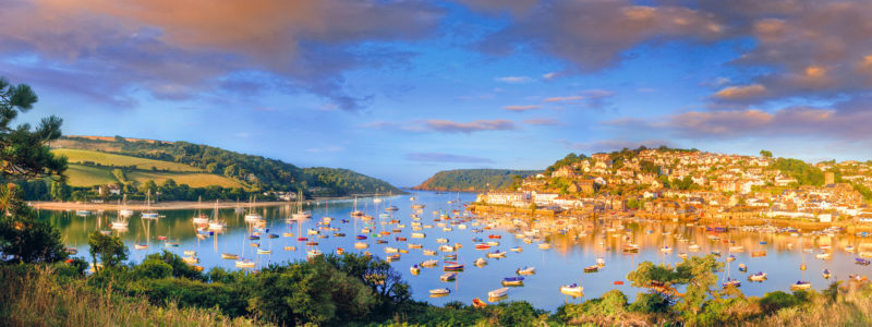 panorama of a wide river estuary filled with fishing boats and yachts with a town on the hill to the right hand side and a grassy hill to the left at sunset with a golden light. best places to visit in south devon england