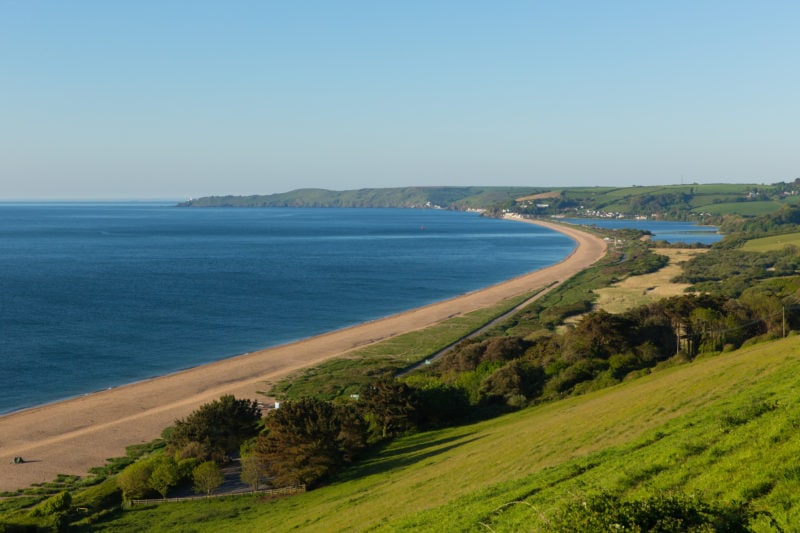 looking down from a grassy hillside towards a large blue bay with a white sany beach and a small headland in the distance - start bay in devon