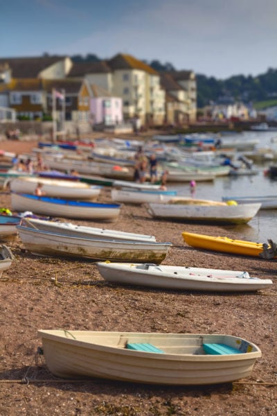 cream and turquoise rowing boat with many more wooden rowing boats behind it on redish shingle sand at teignmouth beach in south devon. the town of teignmouth is out of focus in the background with some white and yellow buildings just visible.