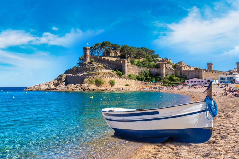 A blue and white rowing boat on a sandy beach with turquoise sea and a hewadland in the background with castle walls and a turret standing amongst the greenery