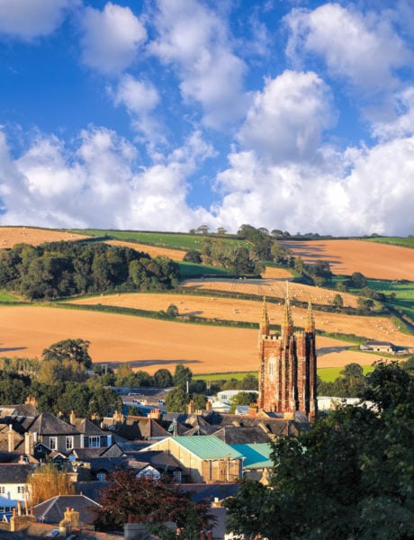 red brick church tower rising out of a small english town in totnes with yellow fields and green hedgerows in the background on a very sunny day in devon with blue sky overhead