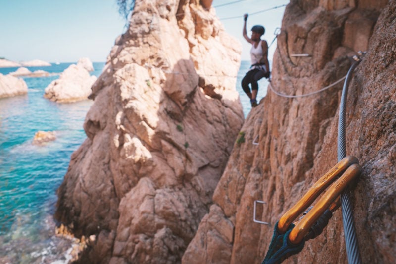 Girl out of focus in the background doing a via ferrata on the coast of catalonia. she is wearing a white t shirt and a helmet but no detail is visible. she is climbing on the edge of an orange cliff with the sea underneath. in focus in the foreground is a metal carabiner clip and the iron wire of the via ferrata Cala de Molí in Sant Feliu de Guixols Spain.
