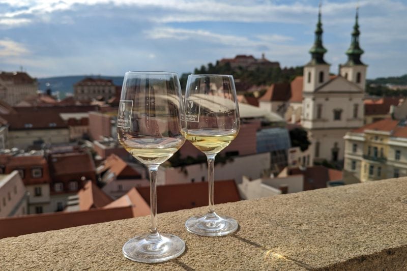 two wine glasses with white wine on a stone wall above Brno town centre with a white two towered building out of focus in the background on a sunny day. 