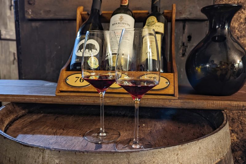 two wine glasses with red wine balanced on top of a wooden barrel in front of three bottles of red wine