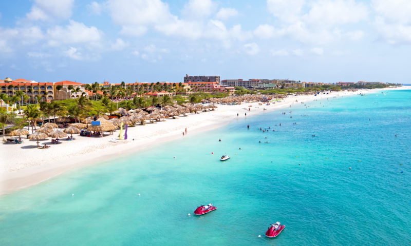 Aerial view of a white sand beach and turquoise sea with some hotels and palm trees along the shoreline and two red jet skis in the water - reasons to visit aruba