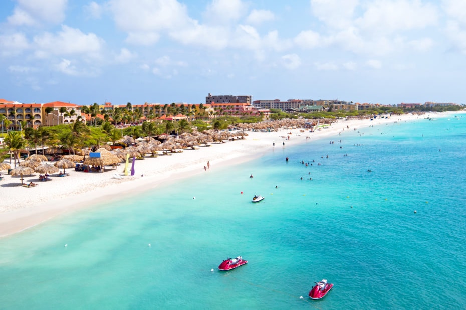 Aerial view of a white sand beach and turquoise sea with some hotels and palm trees along the shoreline and two red jet skis in the water - reasons to visit aruba