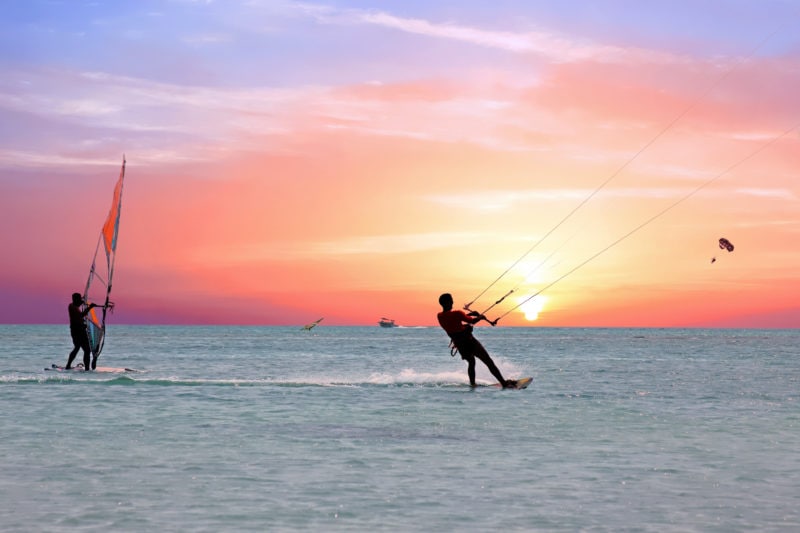 silhouettes of a windsurfer and kitesurfer on flat calm water at sunset with a pink and orange sky behind