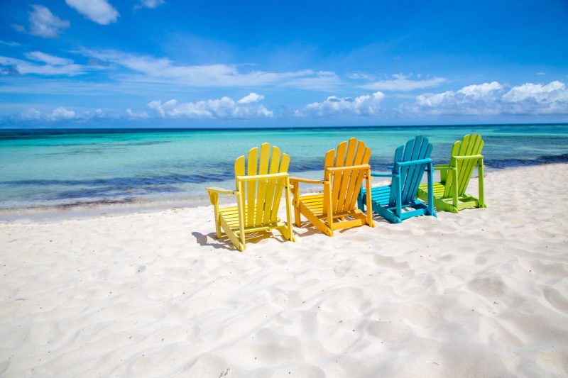 four colourful beach chairs painted yellow, orange, blue and green (left to right) on white sand facing the turquoise ocean. Aruba in the Antilles.