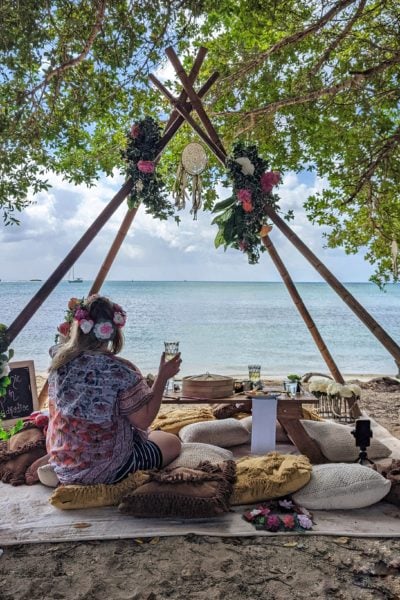 girl wearing a multicoloured shawl and a flower crown sitting facing away from the camera on a beach looking out to sea with a glass of champagne in her hand underneath four wooden poles arranged in a triagular tipi shape with a small table in front and several cusions scattered around