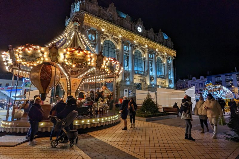 night time scene with an ornate collonaded white building covered in white christmas lights behind a lit up yellow and red merry go round with a few people walking past at calais christmas market. 