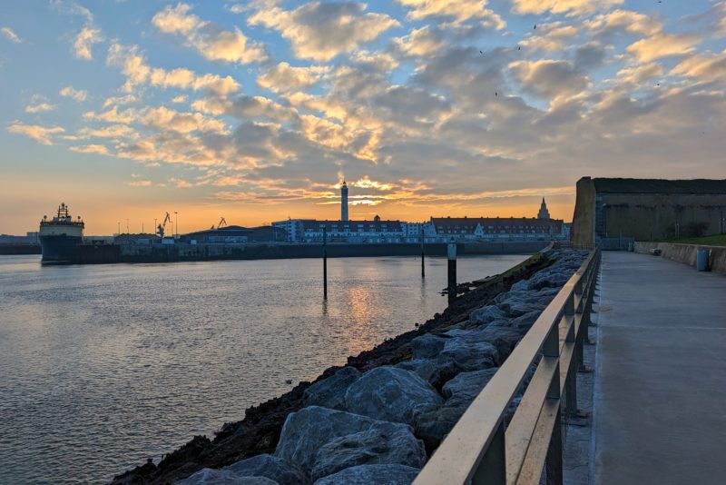 looking along the pier towards Calais with the harbour in the foregound and a sunrise sky behind the town. Calais lighthouse is silhouetted against the orange sky. 