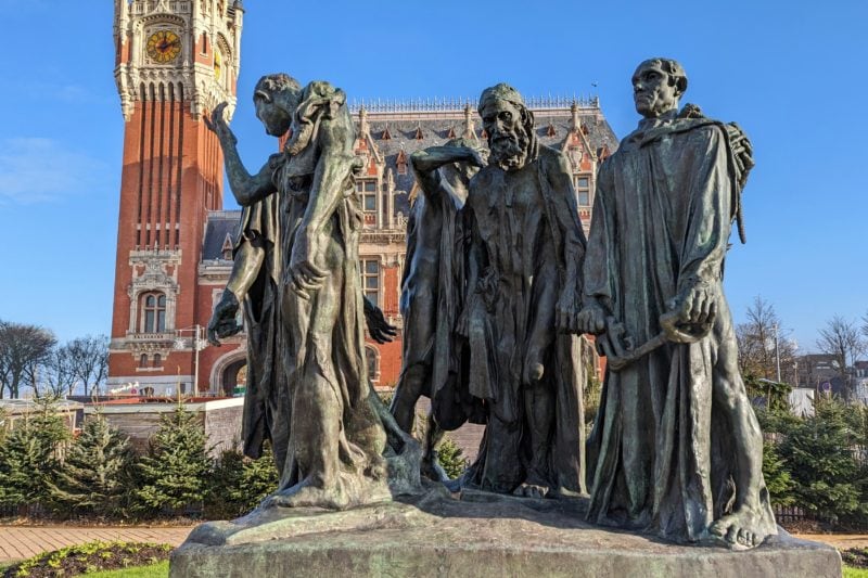 close up of a greenish grey metal statue of five male figures dressed in robes with a red brick town hall with an ornate facade and tall clocktower behind them on a very sunny day in calais with a blue sky overhead. 
