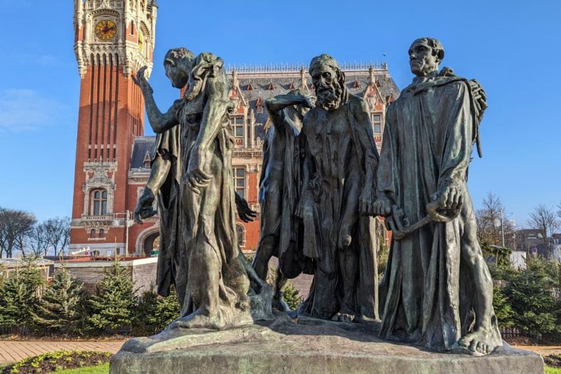 close up of a greenish grey metal statue of five male figures dressed in robes with a red brick town hall with an ornate facade and tall clocktower behind them on a very sunny day in calais with a blue sky overhead.