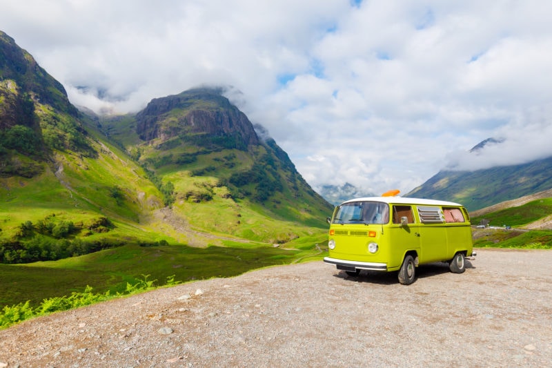 lime green campervan with a white rood on a road with green fields and 3 rocky mountains behind on a sunny day with lots of clouds in the sky. 