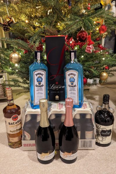 arrangement of bottles in a pyramid shape with two bottles of sparkling wine in front of a large pack of beer bottles with a bottle of rum on either side, two bottles of bombay sapphire gin on top and a black box of Lanson champagne between them and a christmas tree with lights and decorations behind