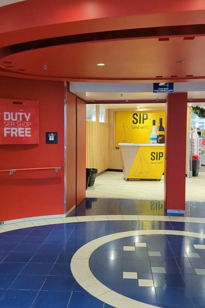 red walls and ceiling over an archway entrance to the DFDS duty free sea shop onboard the ferry. the floor in front of the archway has blue tiles with white tiled circles. inside the archway is a large yellow poster with the words sip and sail. 