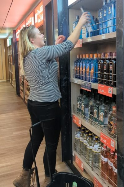 emily wearing black skinny jeans and brown boots and a black and white striped long sleeve top standing in a duty free shop on the DFDS ferry to France in front of shelves of gin bottles reaching down a bottle of bombay sapphire. 