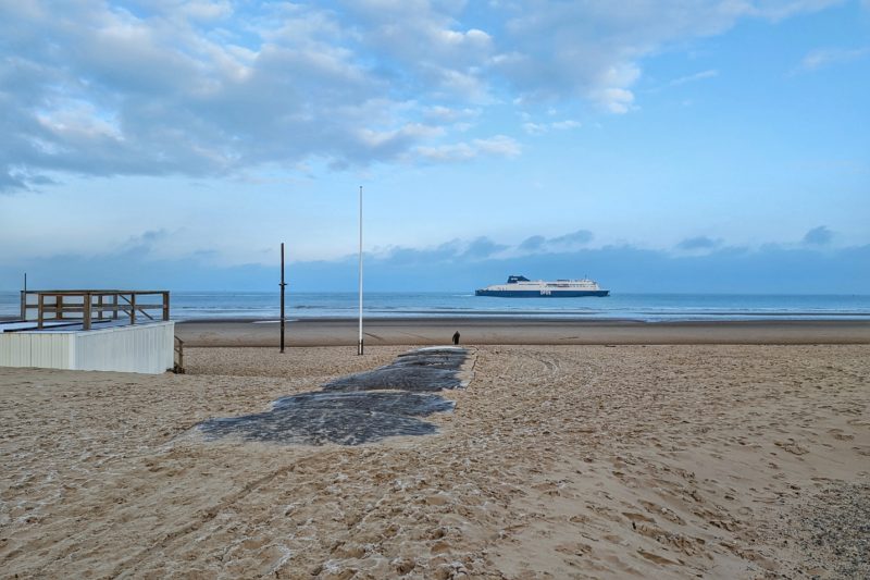 Calais beach on a very cold day in december with frost on the white sand and a DFDS ferry on the blue sea in the background beneath a mostly clear blue sky. 