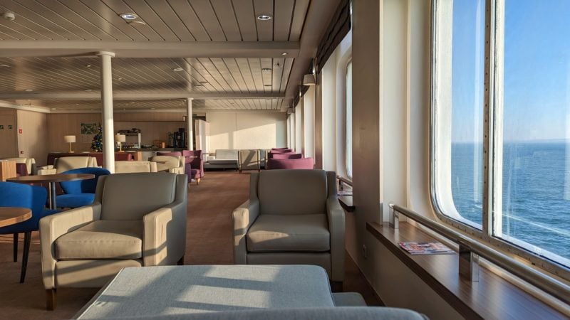 a large empty lounge area on a ferry on a sunny day with blue sea and blue sky visible out of the windows on the right. the lounge has a light brown carpet and cream walls and cieling and is fillwed with cream and burgundy leather armchairs.