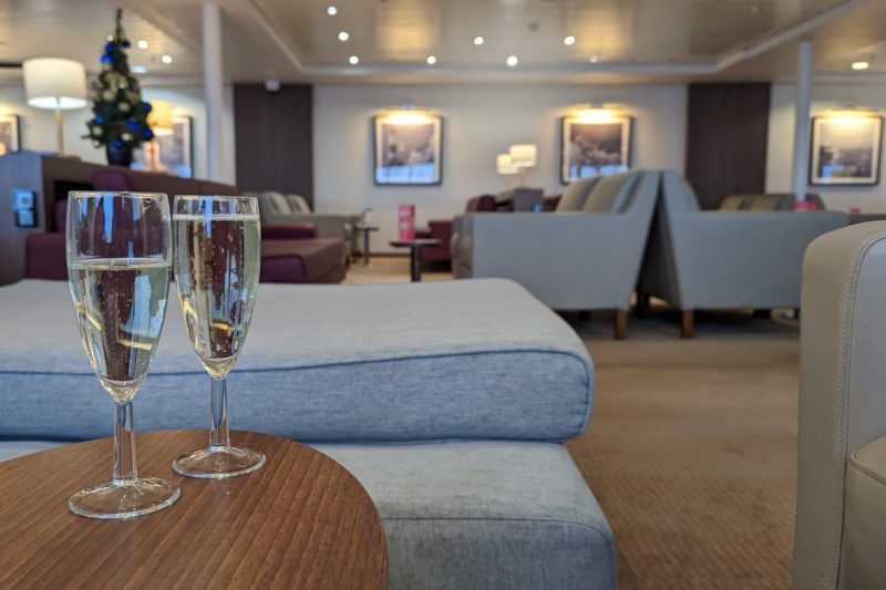 wooden table with two champagne glasses filled with prosecco with a grey pouffe behind and the large DFDS Premium Lounge behind that filled with grey and burgundy leather sofas with cream walls. 