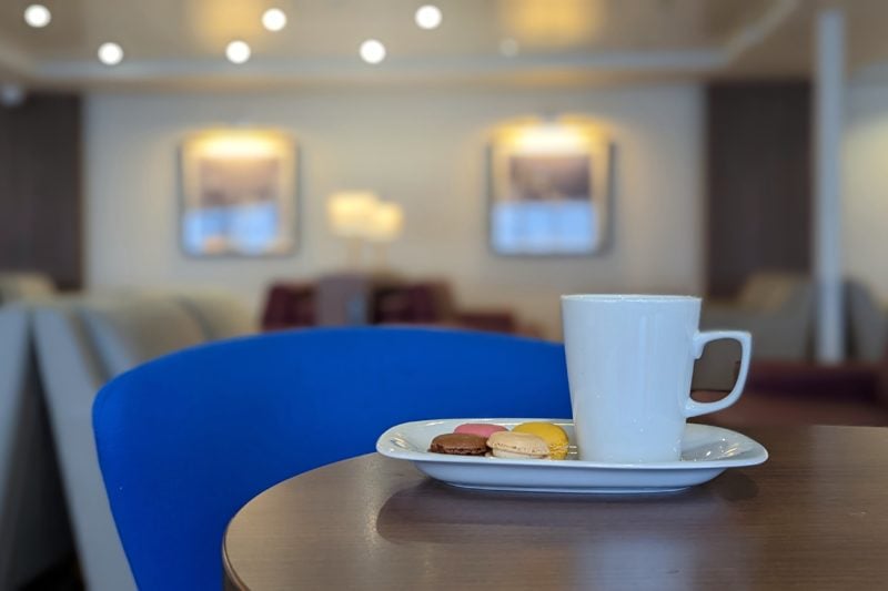 close up of a white mug on a plate with four macarons. there is a bright bluechair at the table and the background is out of focus but shows cream sofas and a cream wall in the DFDS Premium Lounge. 