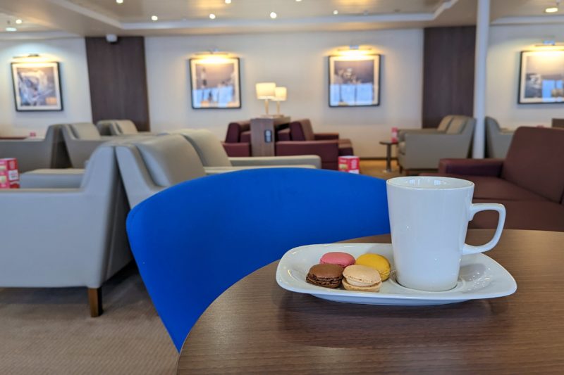 close up of a white mug on a plate with four macarons. there is a bright bluechair at the table and the background is out of focus but shows cream sofas and a cream wall in the DFDS Premium Lounge.