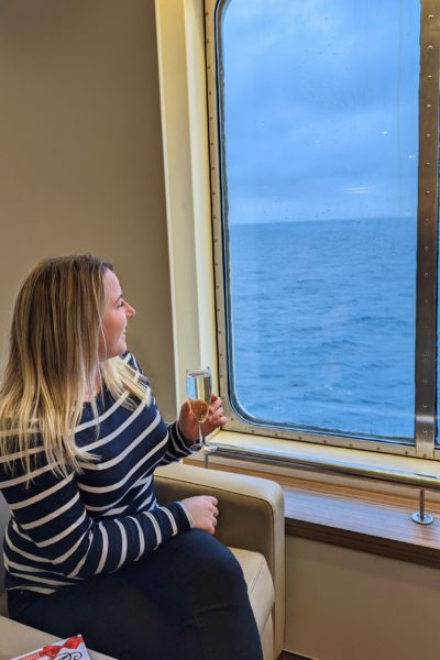 emily sitting in a cream leather armchair by a window on a ferry wearing black jeans and a navy blue and white striped long sleave top holding a glass of prosecco and looking out the window with her long blonde hair down.