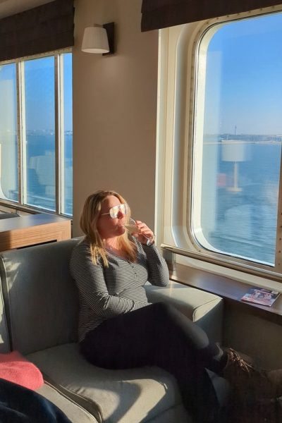 emily sitting on a grey sofa next to a window on a ferry with blue sea visible out the window on a very sunny day. She is wearing black jeans, a white and black striped long sleeved top, and gold sunglasses which are reflecting the sunlight while she sips prosecco and looks out the window with her blonde hair down around her shoulders. 