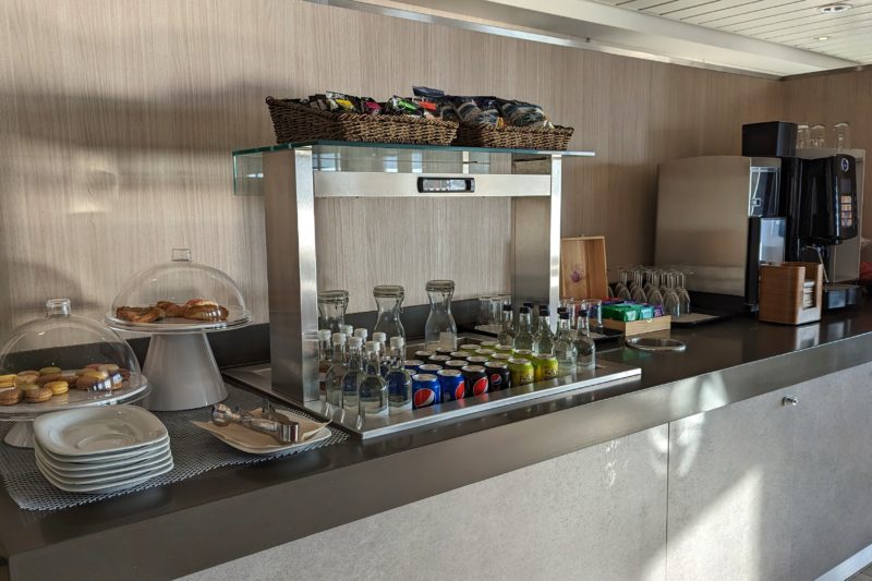 a long grey counter with self serve snacks and drinks, from left to right there is a white cake stand with glass lid containing several colourful macarons, another white cake stand with pastries, a large chilled section full of glass water bottles and canned soft drinks, a wooden box with assorted tea sachets and a large electronic coffee machine
