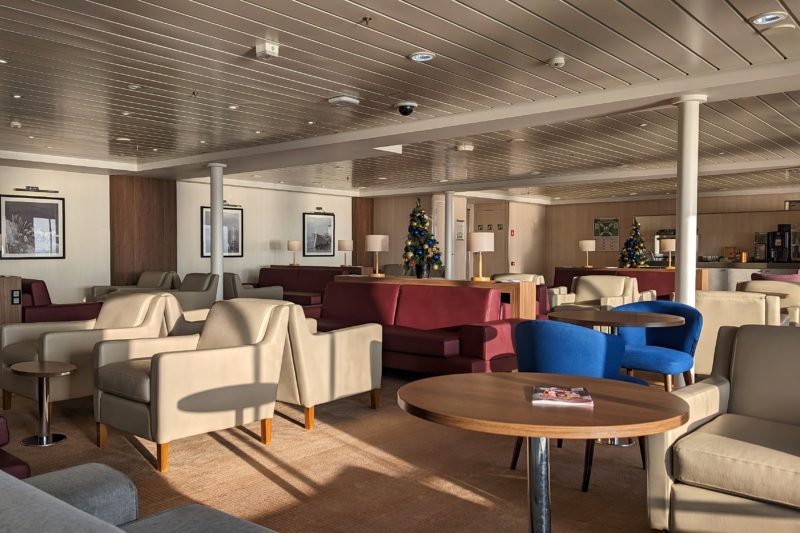 the DFDS Premium Lounge on a sunny day with light brown carpet and cream walls and cieling and multiple cream and burgundy leather sofas and armchairs scattered about. there is a circular wooden table in the foreground and a couple of small decorated christmas trees on tables in the background. 