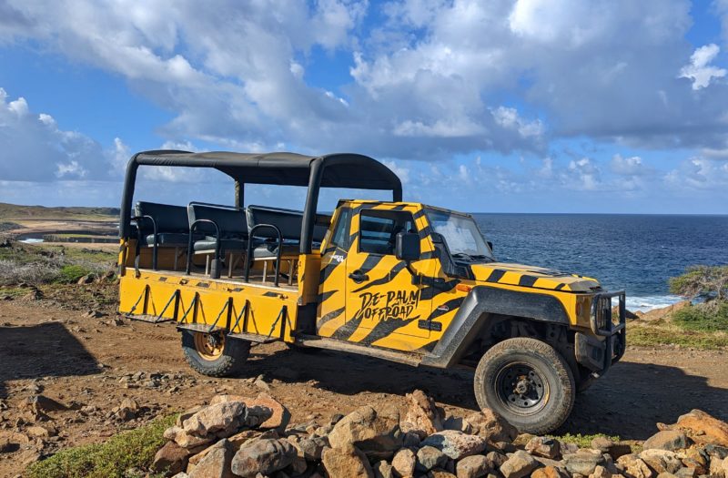 yellow safari jeep with open sides and a black roof parked on orange rocky ground with the blue sea and blue cloudy sky behind it