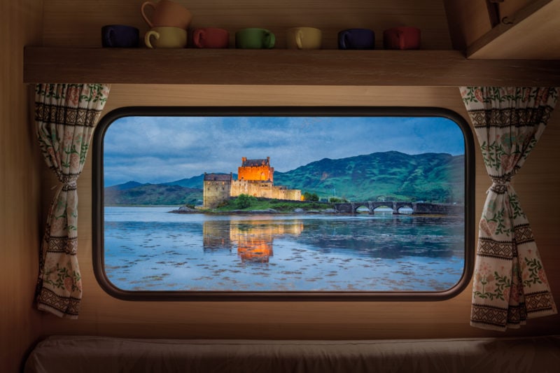 looking through a window with tartan curtains and a shelf of mugs above it with a view of a lake out of the window and a stone castle lit up at dusk with a dark sky and hills in the distance. best places to go in a campervan UK.