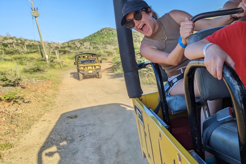 girl in a khaki tank top, cap and sunglasses leaning out of a yellow jeep with a big smile. another yellow yeep is visible on the dirt road behind. fun things to do in aruba.