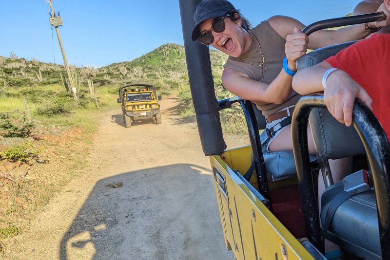 girl in a khaki tank top, cap and sunglasses leaning out of a yellow jeep with a big smile. another yellow yeep is visible on the dirt road behind. reasons to visit aruba. 