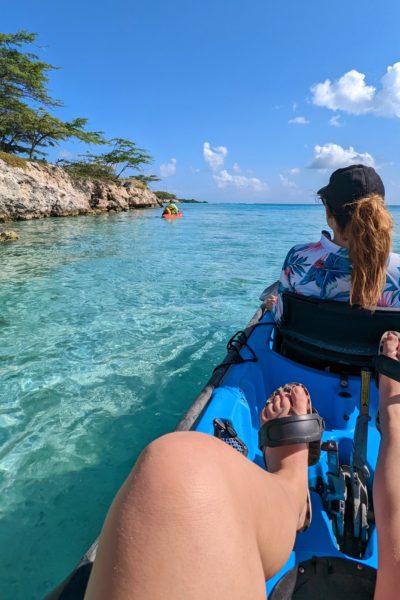 a woman's legs in a bright blue kayak with a girl sitting in front with brown hair in a ponytail and a black cap on looking out at the clear turquoise sea with the white rocks of the shore to the left