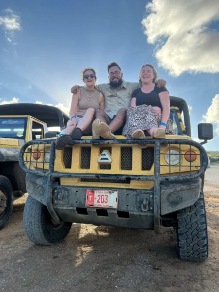 three people sitting on the hood of a yellow jeep with blue sky behind. from left to right is a white girl with brown hair wearing a  beige tank top, an Aruban man with dark hair and beawrd wearing glasses and a beige shirt, and a blonde white girl wearing a black t shirt and leopard print skirt. all three are laughing. 