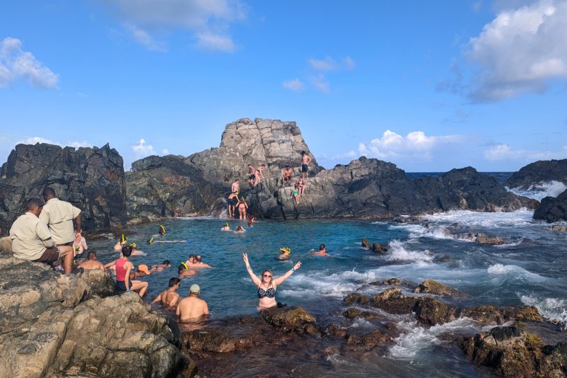 emily wearing a blue polka dot bikini with her arms in the air smiling at the camera. she is standing in a natural rock pool with a number of other people around standing and swimming. there are large grey rocks in the background surrounding the blue pool. unique things to do in aruba. 