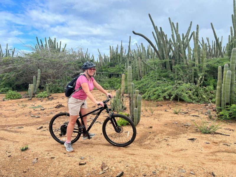 emily in a pink t shirt and beige shorts with a black helmet on riding a black bike on a sandy path with tall green cacti behind her