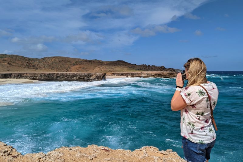 emily wearing denim shorts and a white t shirt covered in green palm trees taking a photo with the bright blue sea behind her and distant orange coloured cliffs behind that