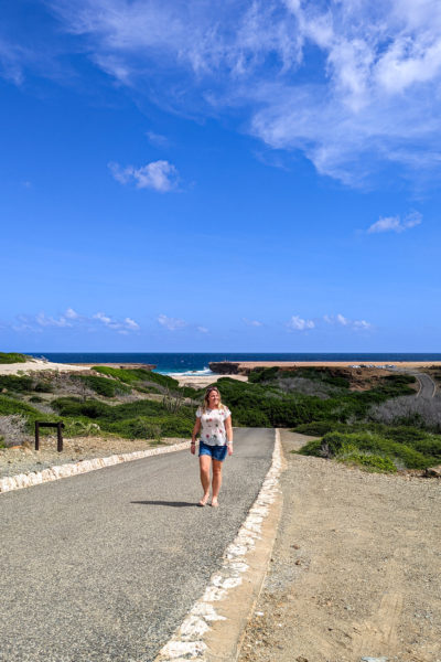 emily wearing denim shorts and a white t shirt covered with green palm trees walking towards the camera up a tarmac road with a green area full of cactuses behind her and the sea behind that on a very sunny day with bright blue sky overhead - fun things to do in aruba
