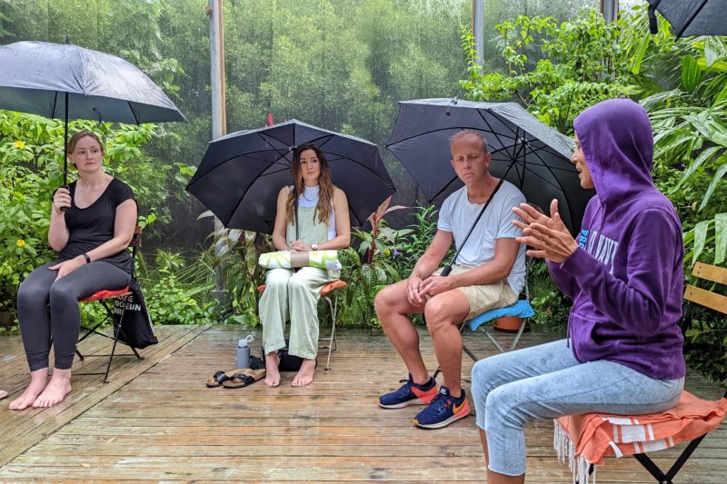 four people sitting in a semi circle on wooden chairs on a wooden deck all holding large black umbrellas, the lady closest to the camera on the right is leading a meditation session and is wearing a bright purple hoody and light blue jeans. it is raining and there is dense green foliage in the background.