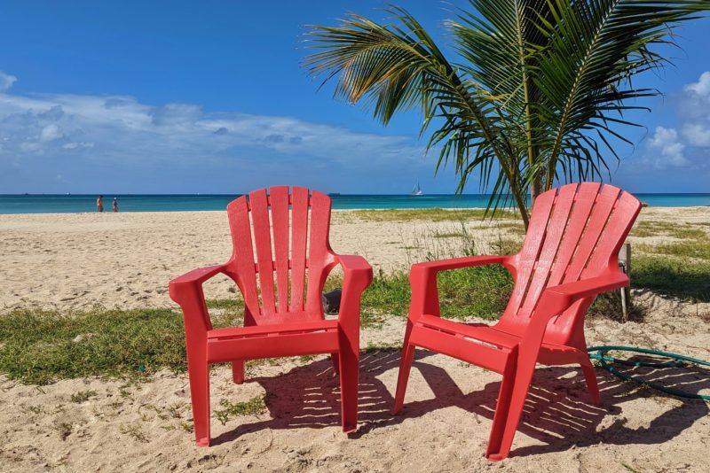 two red plastic lawn chairs on a white sandy beach with a palm tree behind them and the turquoiuse sea behind that on a calm sunny day with blue sky above. reasons to visit Aruba