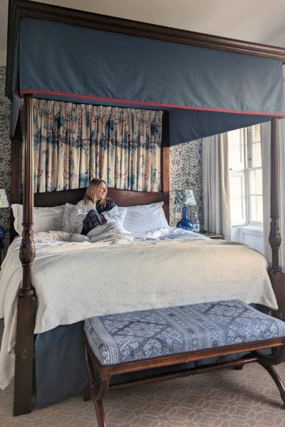 Emily sedang duduk di tempat tidur bertiang empat dengan baju tidur biru tua dan jubah putih, memandang ke luar jendela di sebelah kanan.  Tempat tidurnya memiliki seprai putih dan bingkai kayu gelap dengan bangku kaki berwarna biru di depannya. 
