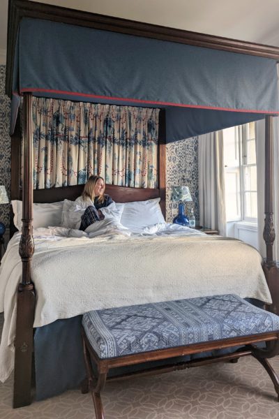 emily is sitting on a four poster bed wearing navy blue pyjamas and a white robe and looking out the window to the right. the bed has white sheets and dark wooden frame with a blue footstool in front of it. 