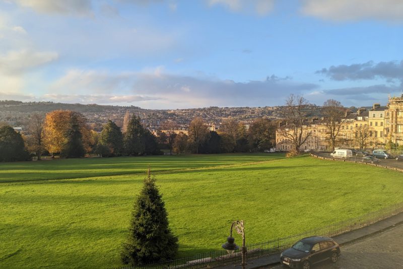 view out a third floor window at the Royal Crescent Hotel and Spa of a large green lawn surrounded by trees with a city just visible in the distance behind and a blue sky overhead on a sunny morning in winter