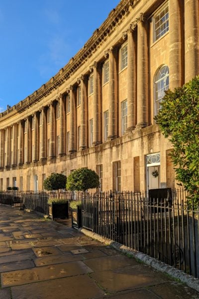 exterior of a three storey georgian townhouse in Bath Englamnd built from beige coloured stone with stone collonades on the upper two floors. there are two small green bushes outside the entrance gate. 